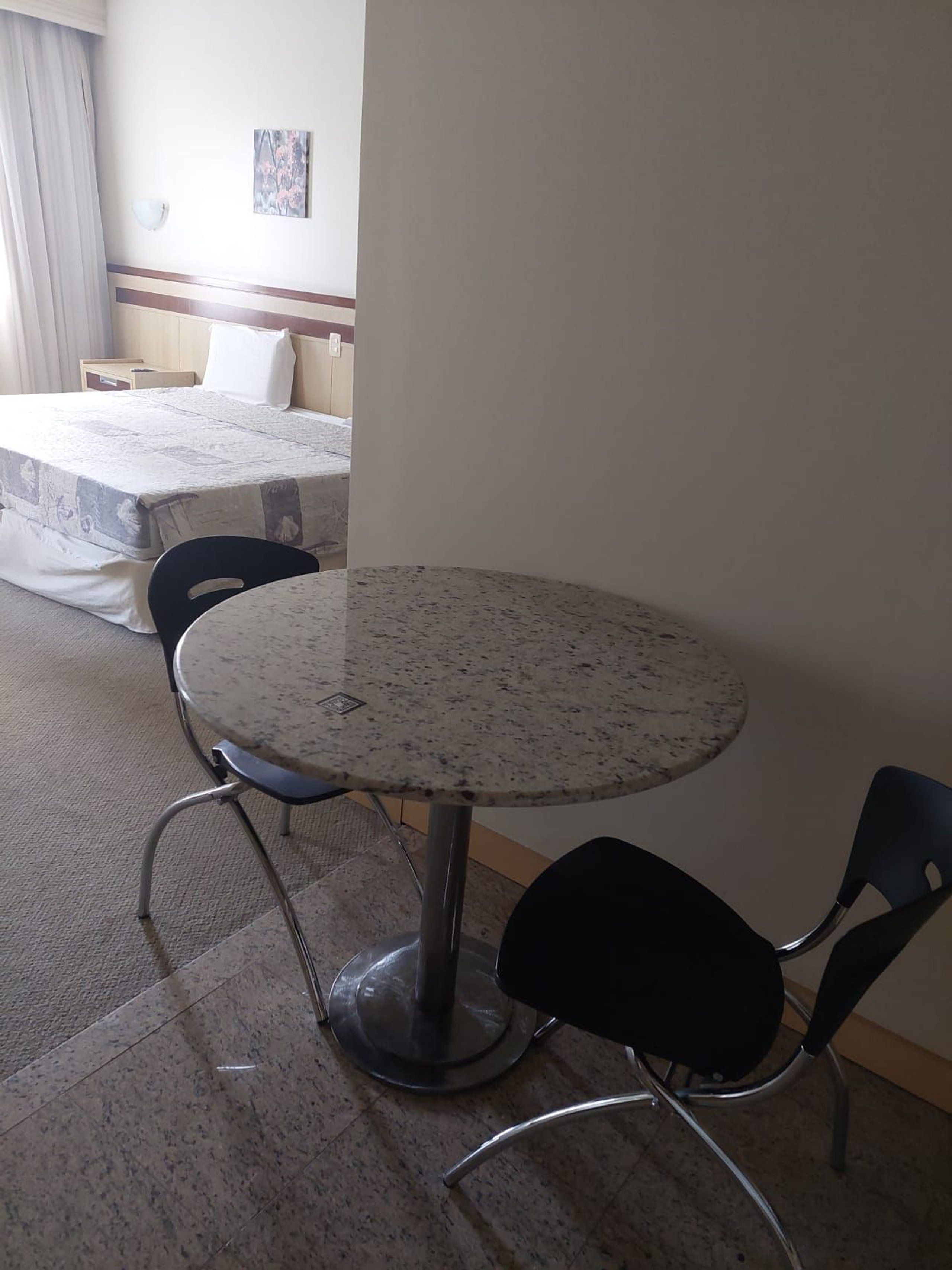 A hotel room with a round table and two chairs, a bed with a white mattress, a nightstand, and a wall-mounted lamp.