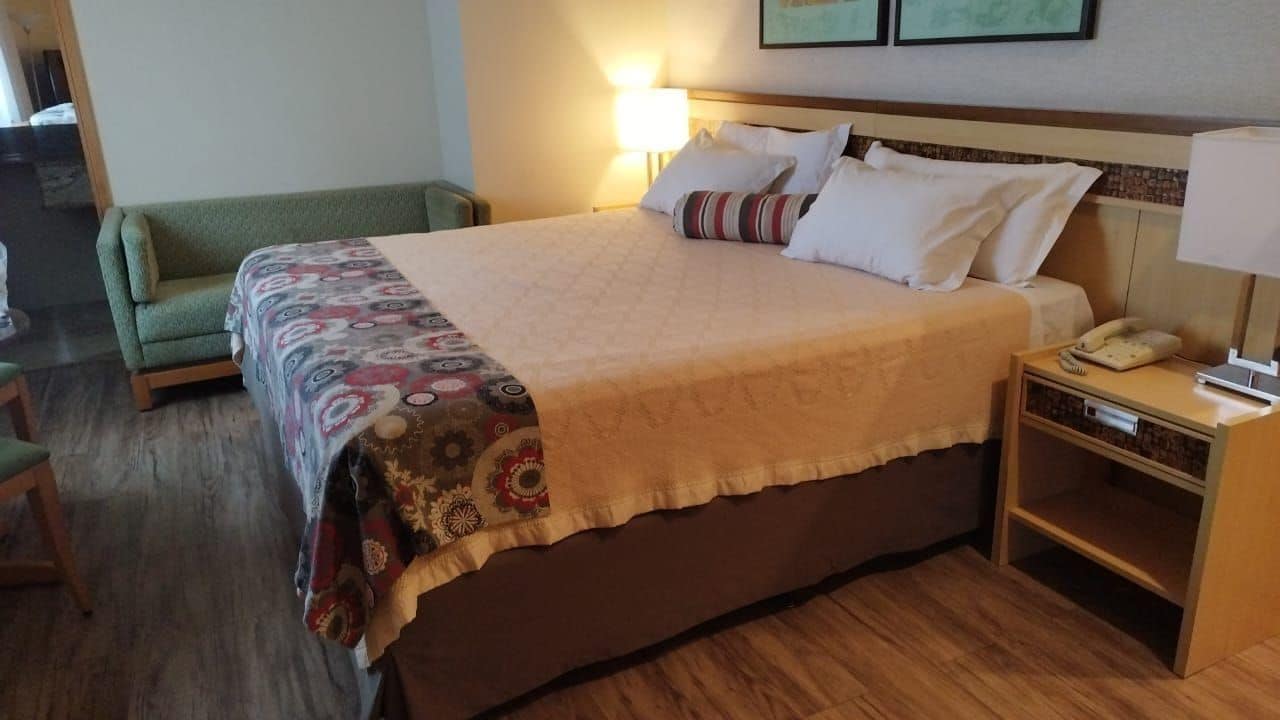 Une chambre d’hôtel avec un grand lit, une table de chevet avec une lampe, un fauteuil vert et un plancher en bois.