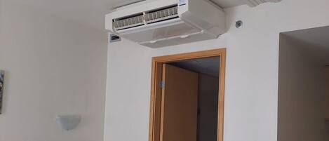A hotel room with a bed, bedside table, a phone, an air conditioning unit, a door, and a small table with a chair.