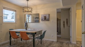 Un coin repas comprenant une table, des chaises et un lustre. On y trouve une fenêtre avec des stores, un miroir et un tableau accroché au mur.