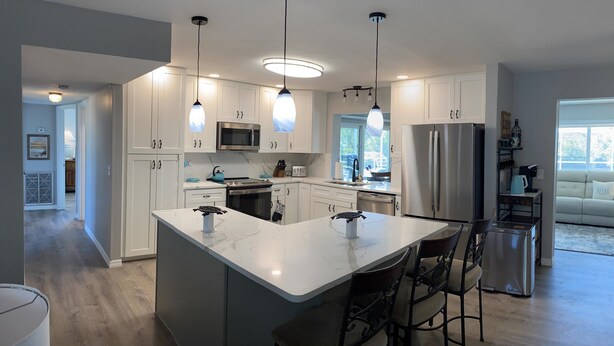A modern kitchen with white cabinetry, a central island with bar stools, stainless steel appliances, pendant lighting, and a view to a living area. - Serenity now, full refund*, pool heat, sleeps 8 (Rotonda West)