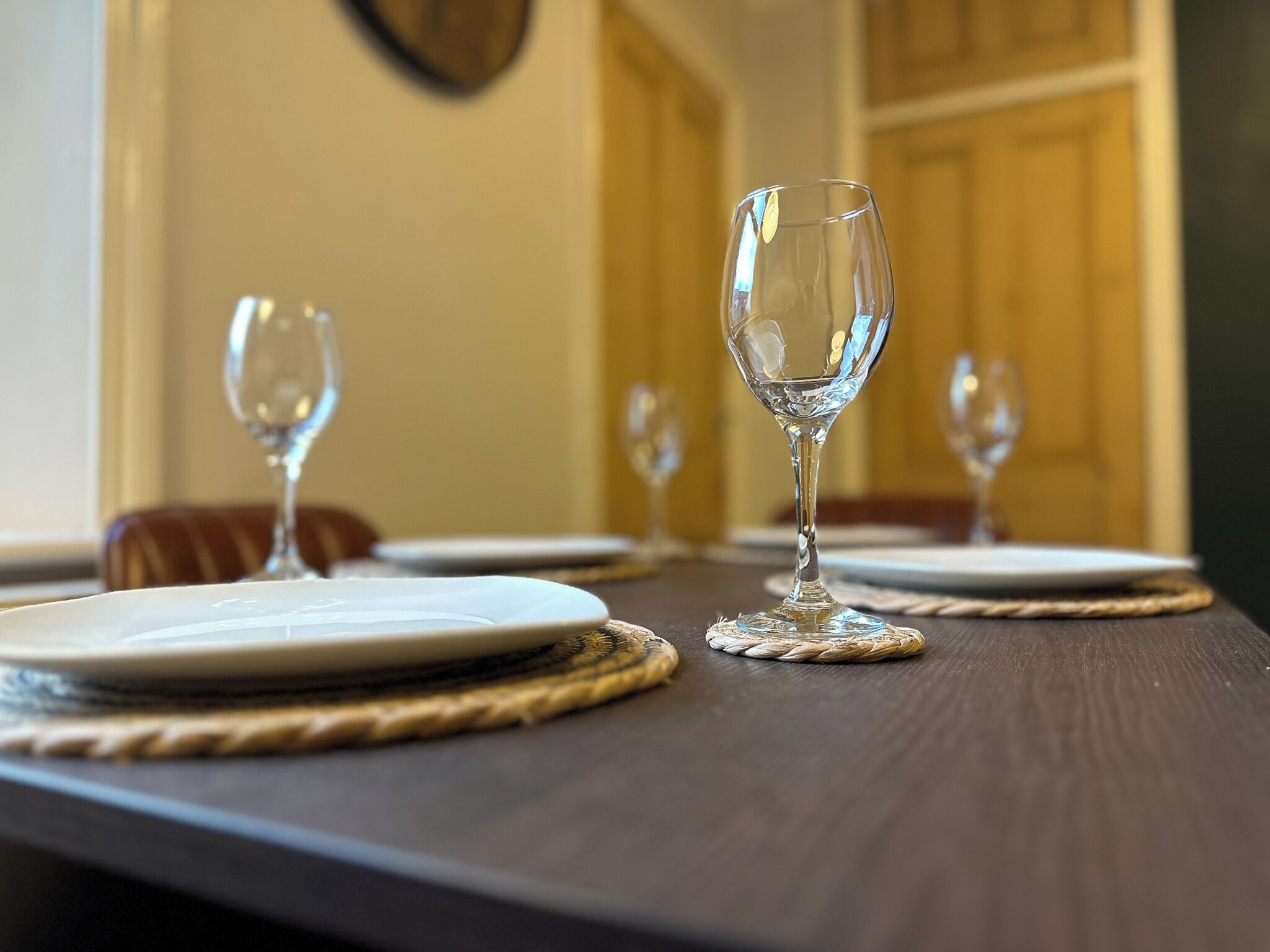 Mesa de jantar posta com três copos de vinho vazios e pratos, adornada com jogos americanos de palha.