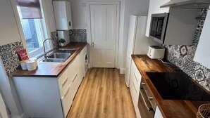 Une cuisine compacte avec un plancher en bois, des meubles blancs, un four à micro-ondes encastré et une fenêtre avec des stores.