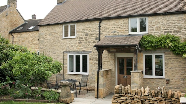 Une maison en pierre avec un toit en tuiles, un petit jardin entouré d’un muret, et une terrasse aménagée avec du mobilier d’extérieur.