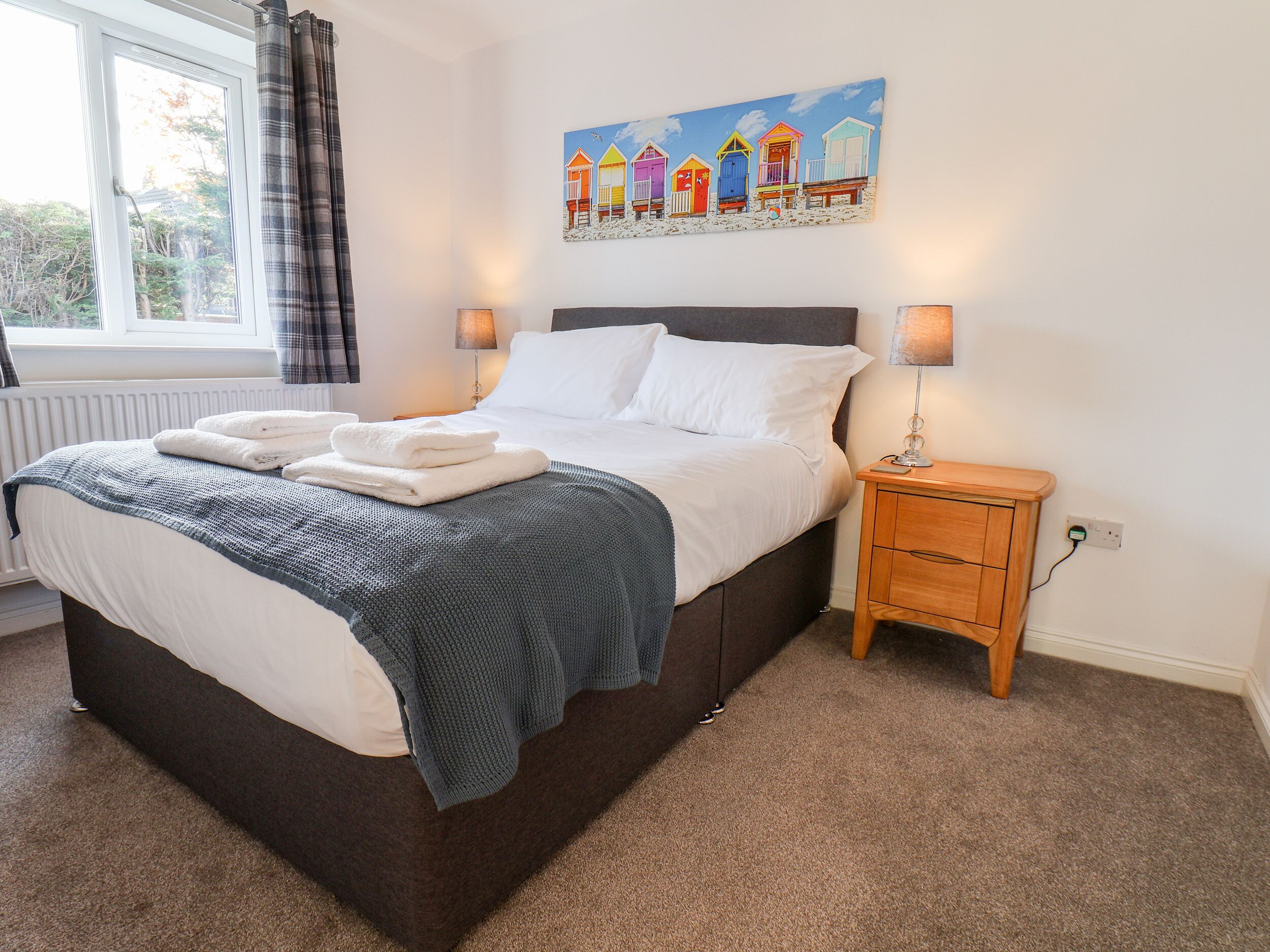 A neatly made bed with white linens and a blue blanket, a bedside table with a lamp, and a painting of colorful beach huts on the wall.