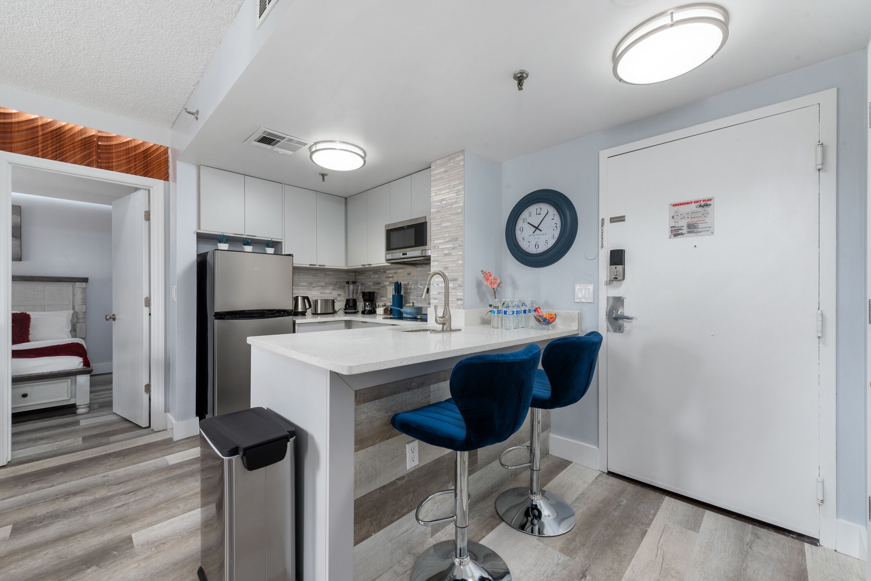 Een moderne keuken met witte kasten, een roestvrijstalen koelkast en een magnetron. Er staan twee blauwe barkrukken bij het eiland en een klok hangt aan de muur.