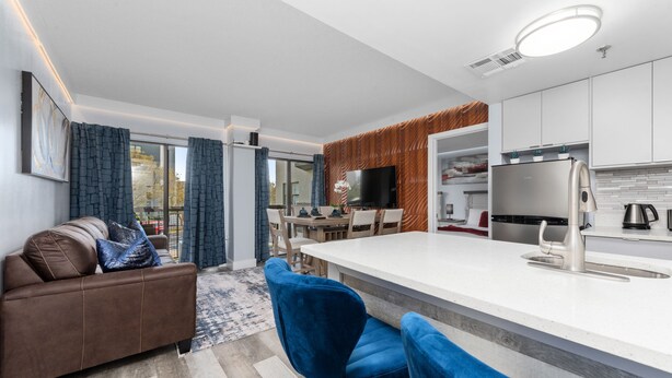 Une cuisine moderne avec un îlot central, un coin repas avec une table et des chaises, un canapé avec des coussins et une grande fenêtre avec des rideaux.