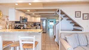 A kitchen with white cabinets, a built-in microwave, and a stainless steel refrigerator. There is a dining area with a white table and chairs. A staircase leads to an upper level. - Beautiful Direct Ocean Front Townhome in Sea Village with Relaxing Ocean Views! (Ocean City)