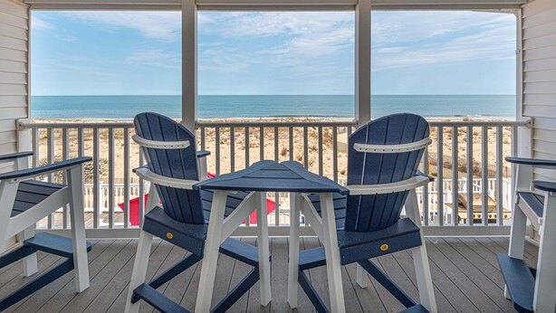 A covered deck with a table and two Adirondack chairs, overlooking a beach. - Beautiful Direct Ocean Front Townhome in Sea Village with Relaxing Ocean Views! (Ocean City)