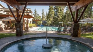 A hot tub with a wooden deck, surrounded by lounge chairs and umbrellas, with a pool and trees in the background. - Sleeps 8 • 3BR • Lux Northstar • Free Shuttle • Fireplace • Xbox • Gym • Hot Tub (Truckee)