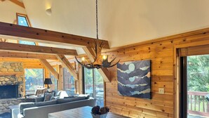 A cozy cabin interior with a stone fireplace, wooden beams, a large dining table, and a scenic view through the windows. - Sleeps 8 • 3BR • Lux Northstar • Free Shuttle • Fireplace • Xbox • Gym • Hot Tub (Truckee)
