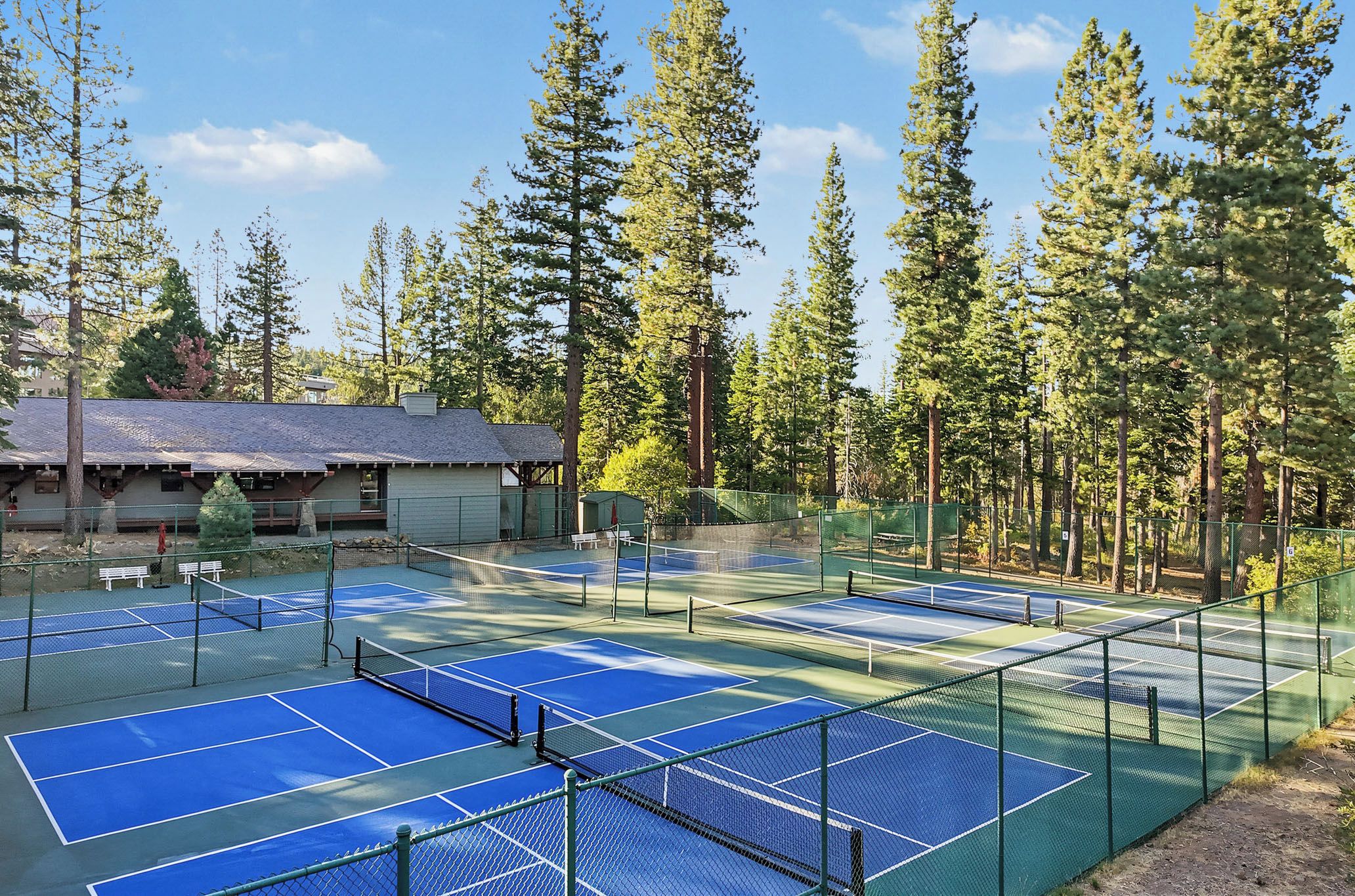 En utomhus tennisbana med flera banor, omgiven av höga träd och en byggnad med ett sluttande tak.