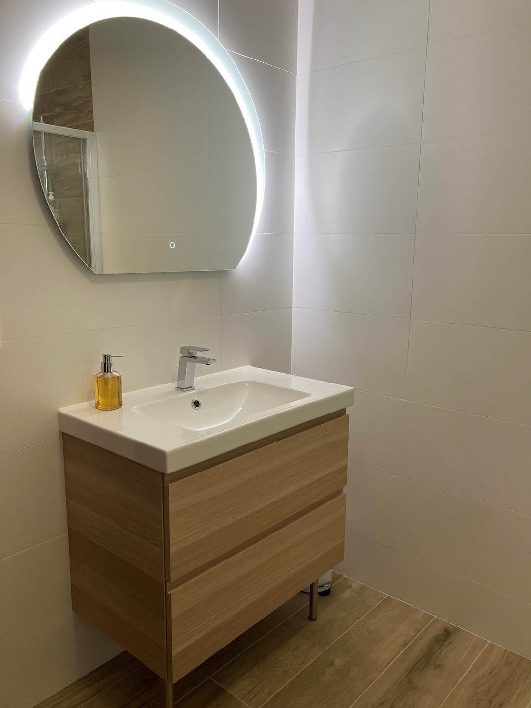 Een moderne badkamer met een witte wastafel, een houten opbergmeubel en een ronde spiegel met verlichting.