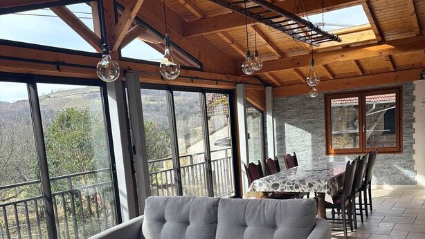A cozy room with a grey sofa, a wooden coffee table, and a dining table with chairs. Large windows offer a view of the outdoors.