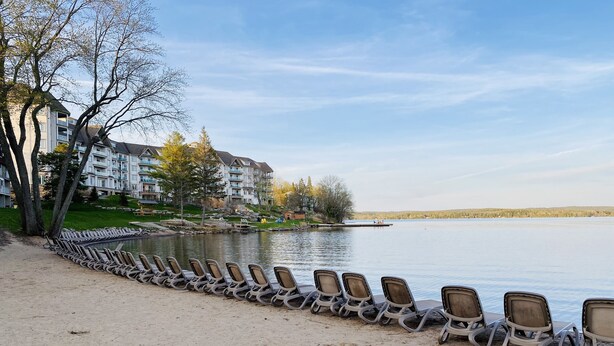 Une vue sur le lac, avec une rangée de chaises longues vides le long de la rive, un bâtiment doté de nombreux balcons en arrière-plan et un ciel dégagé.