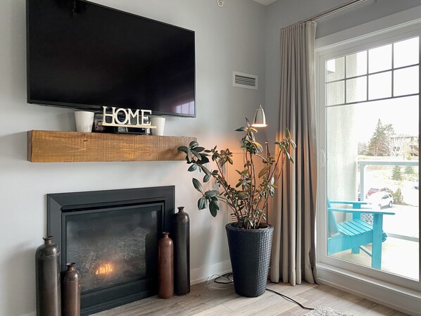 A living room with a fireplace, a TV mounted on the wall, a potted plant, and a window with a view of the outside.