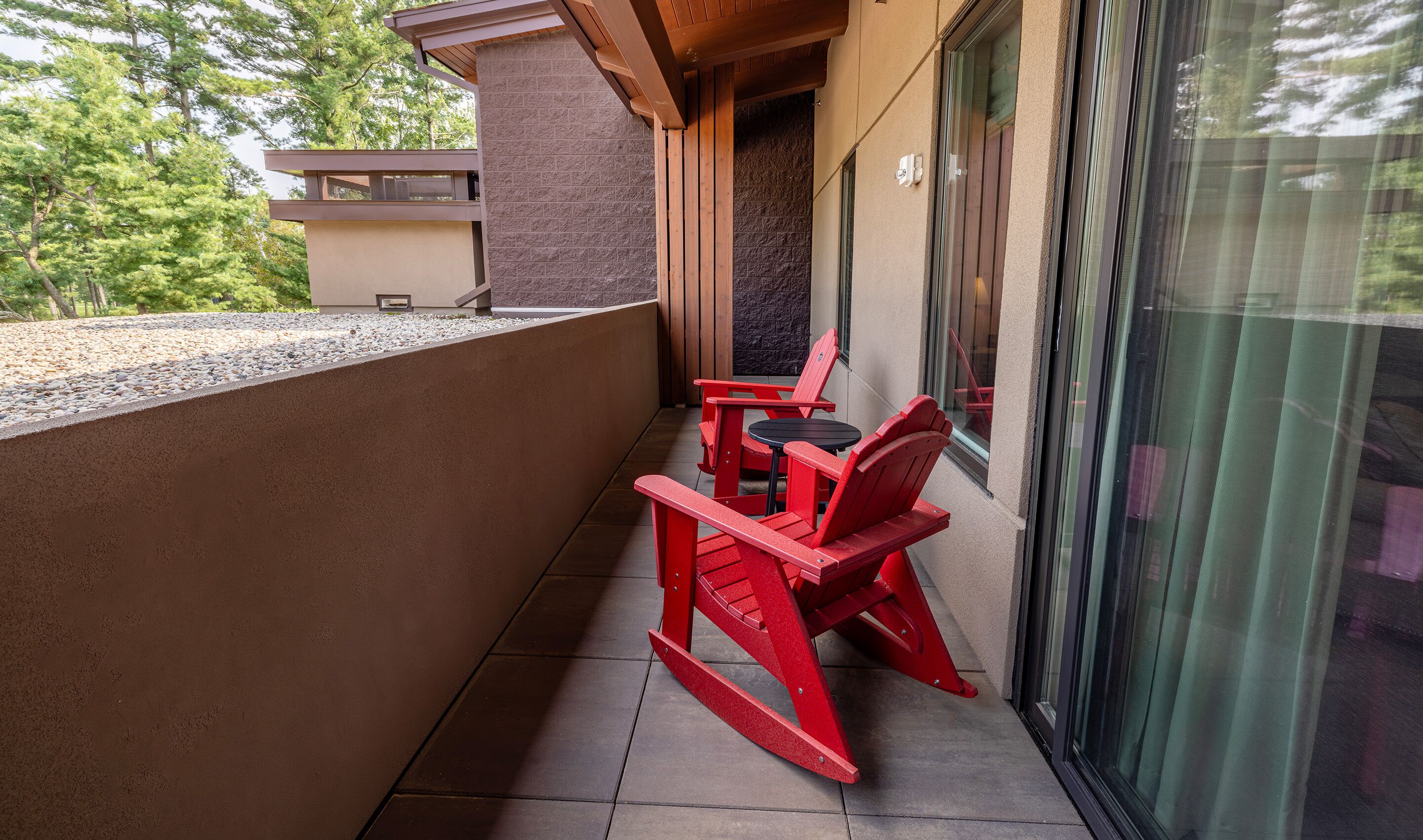 A balcony with a red Adirondack chair and a small round table, a stone wall, and a view of trees.