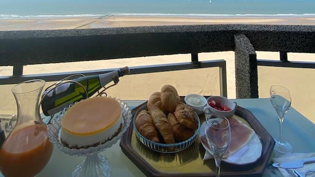 A table with croissants, a cake, and a glass of juice, overlooking a beach. - Amazing 2-bedroom Apartment With Amazing Sea-view (Ostend)