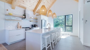 Una cocina moderna con armarios blancos, una isla central con taburetes y vigas de madera en el techo, y ventanas amplias con vistas a la vegetación.