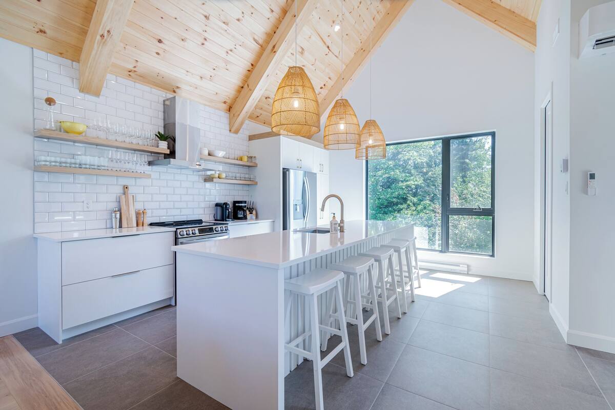 A modern kitchen with white cabinetry, a central island with bar stools, wooden ceiling beams, and large windows offering a view of greenery.