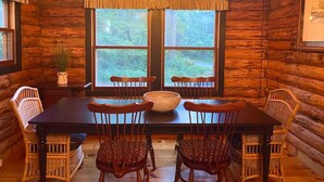 Ein Holzblockbau-Innenraum mit einem Esstisch, vier Stühlen, einer Chandelüster und einem großen Fenster mit Blick auf einen Wald.