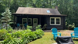 Eine Blockhütte mit Veranda, zwei blaue Adirondack-Stühle und ein überdachter Grill.