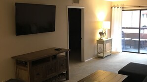 Un soggiorno con TV a schermo piatto, un mobile TV in legno, un divano, un tavolino da caffè e una porta scorrevole in vetro che dà su una zona esterna.