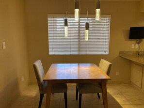 Un coin repas comprenant une table rectangulaire et quatre chaises, une fenêtre avec des stores et des luminaires suspendus au-dessus.