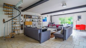 En rummelig opholdsstue med en moderne trappe, en grå sofasæt, en rød lænestol og et stort vindue med udsigt over grøn natur.