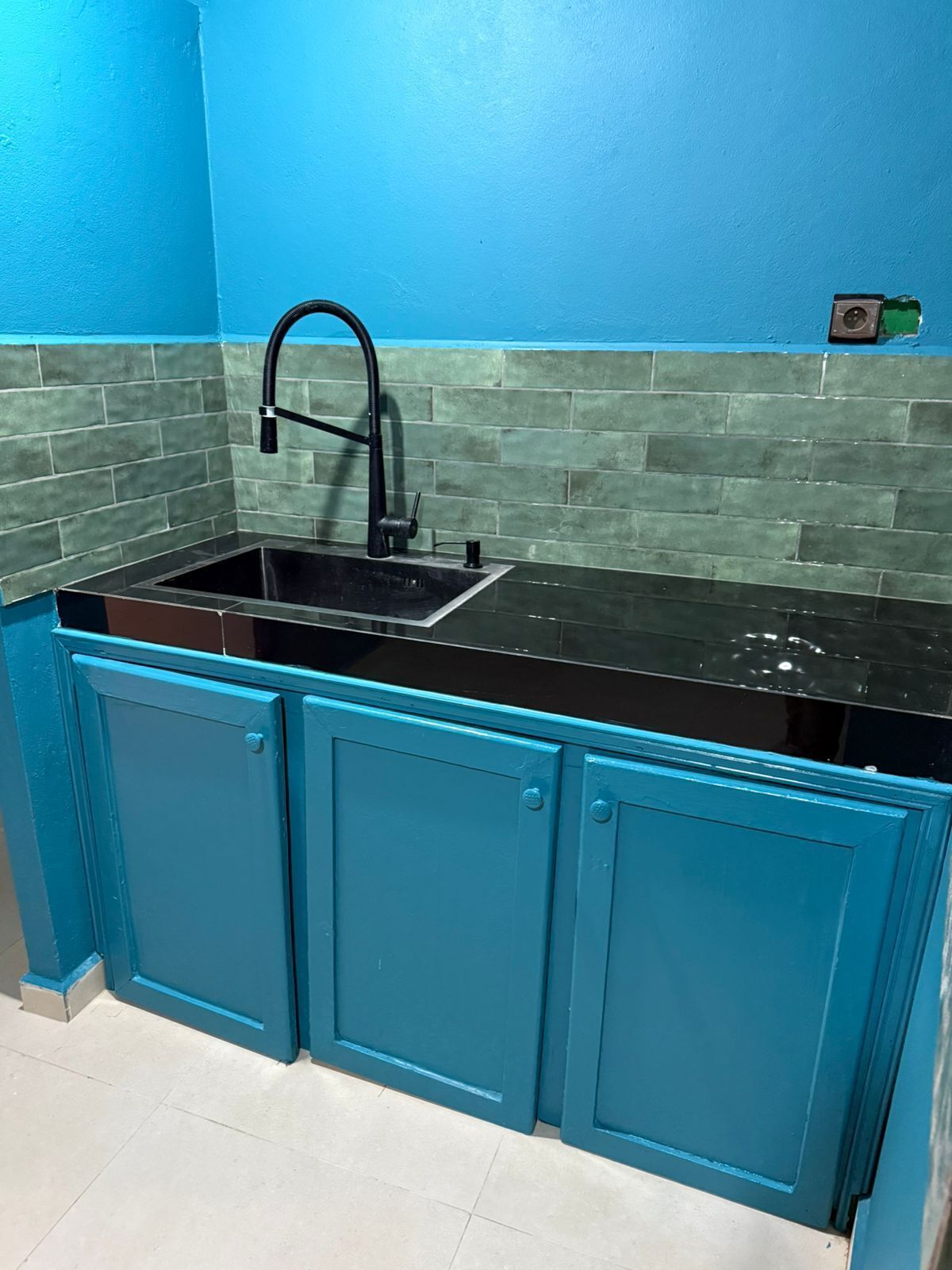 Una cocina con gabinetes azul verdoso, una encimera negra y un lavamanos con grifo negro.