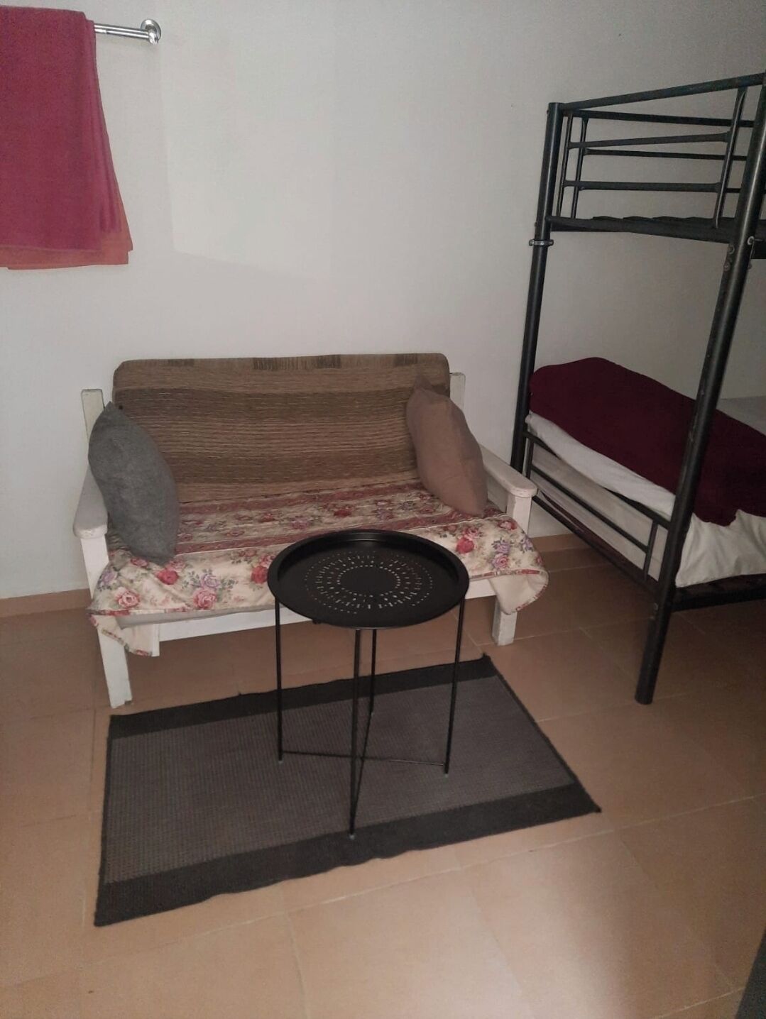 En køjeseng med madras, en sofa med blomstret betræk, et lille rundt bord og et rødt håndklæde på en metalreol.