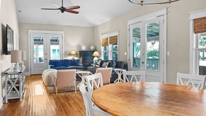A spacious living room with a wooden dining table, white chairs, a blue sofa, and a ceiling fan.