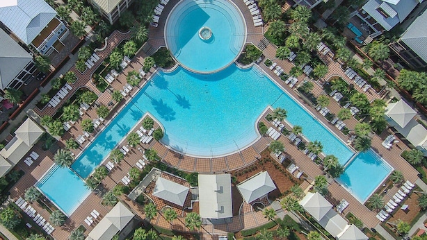 An aerial view of a hotel with a central circular pool surrounded by lounge areas and palm trees.