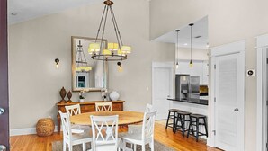 A dining area with a wooden table and white chairs, a chandelier, and a kitchen with white cabinets and a refrigerator.