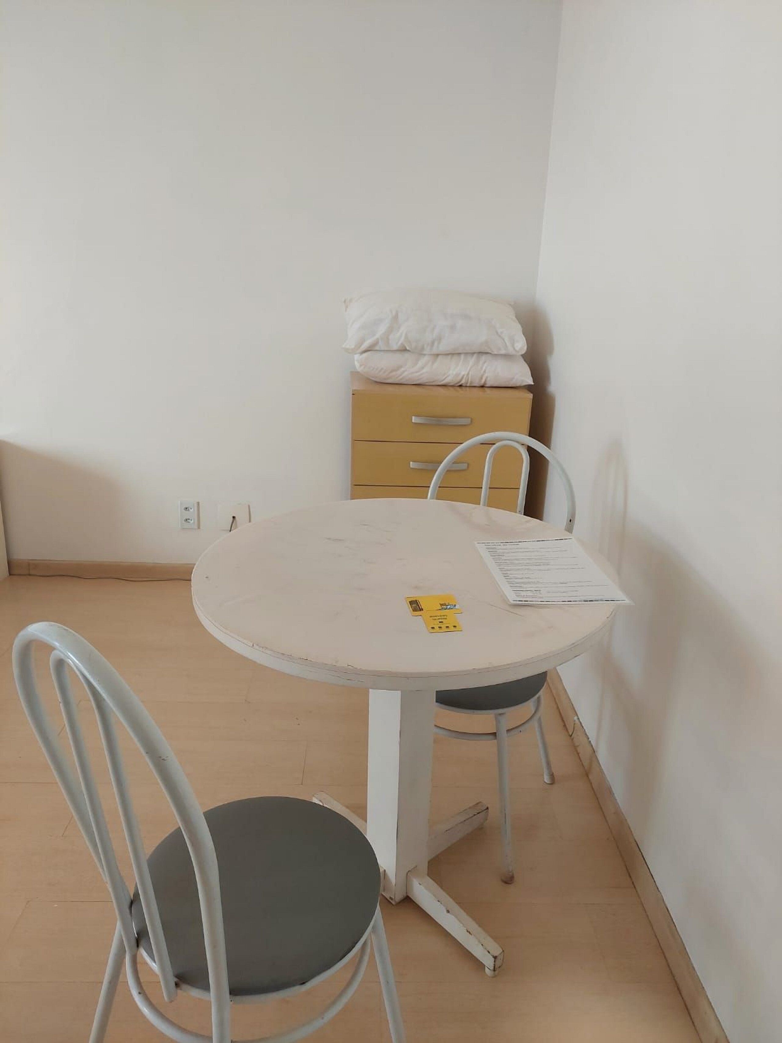 A small, round table with a white surface and a single chair. There is a yellow card and a piece of paper on the table. A beige cabinet with a drawer is in the background.