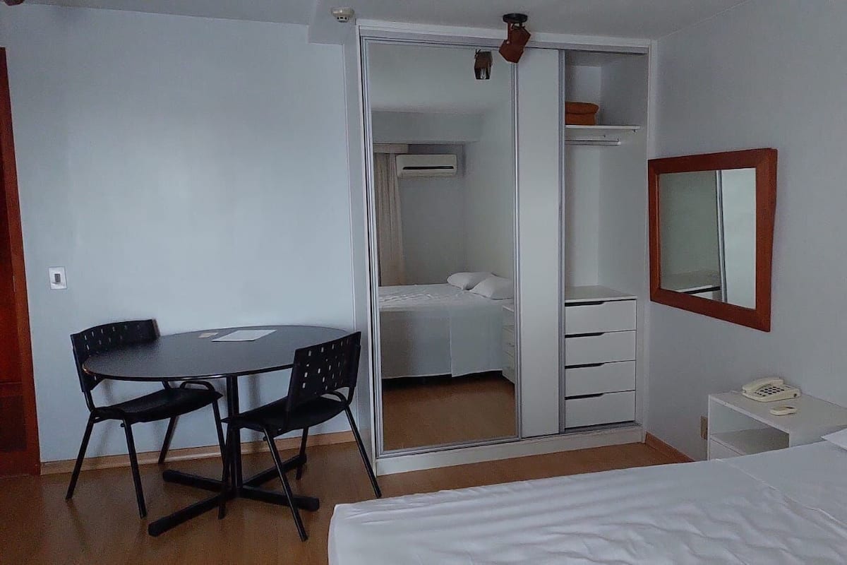 A bedroom with a bed, a desk with chairs, a wardrobe, and a bedside telephone.