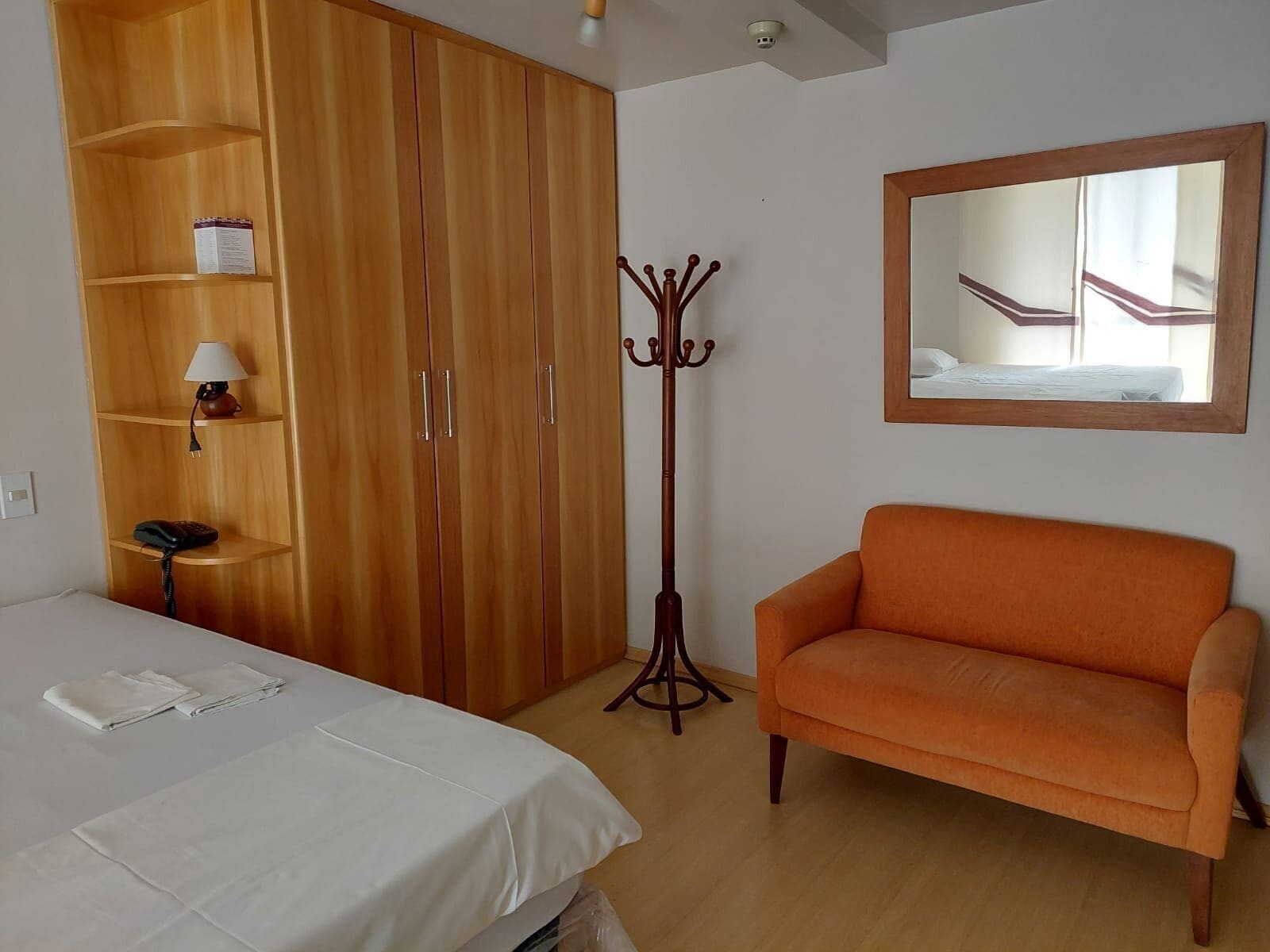 Une pièce comprenant un lit, une armoire en bois, un petit canapé orange, une lampe sur pied et un miroir au mur.