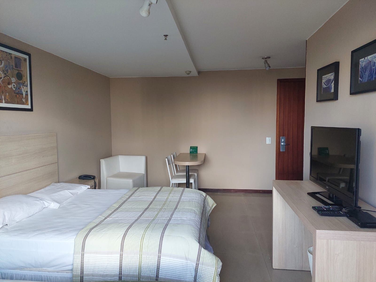 A hotel room with a bed, a desk with a TV, a chair, and a small table.