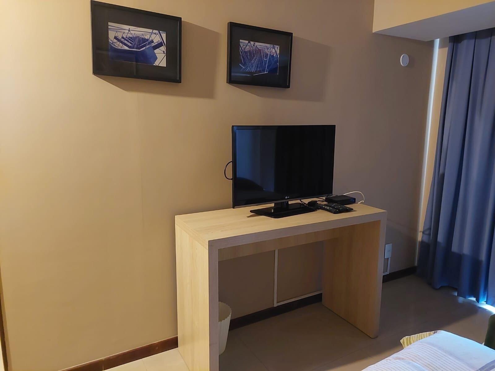 A hotel room with a TV on a wooden desk, two framed pictures on the wall, and a window with curtains.