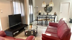 Ein Wohnzimmer mit Flachbildfernseher, rotem Ledersofa, einem Glastisch mit Topfpflanzen, einem Essbereich mit schwarzem Metalltisch und -stühlen sowie einer Wanduhr.