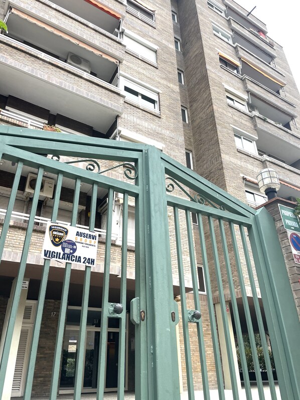 Entrada protegida por un portón a un edificio, con un cartel de seguridad, aviso de vigilancia las 24 horas y letrero de propiedad privada.