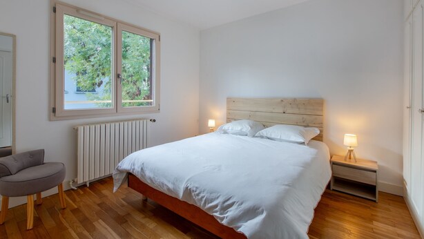A bedroom with a wooden headboard, a bed with white bedding, a nightstand with a lamp, a radiator, and a window with a view of greenery.
