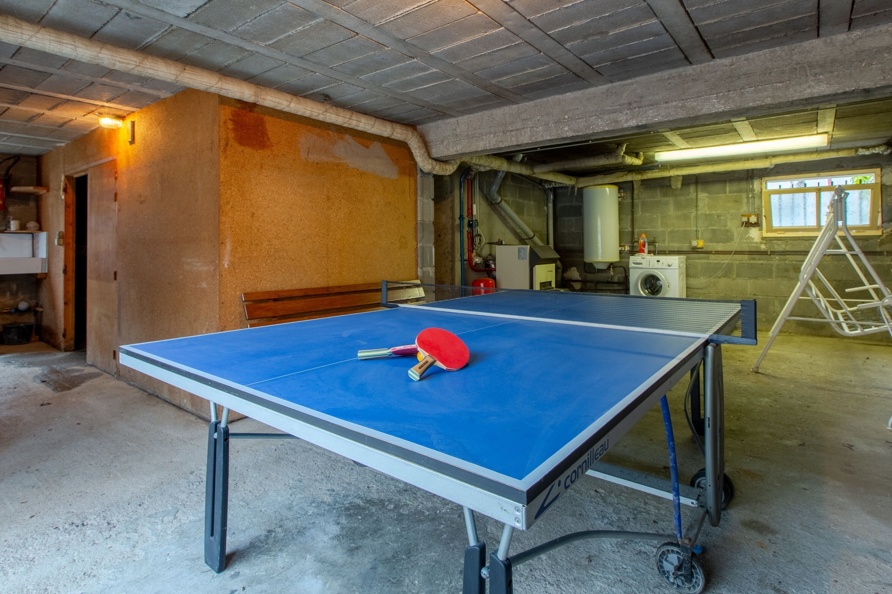 Une table de ping-pong dans une pièce dont le sol est en béton, avec une échelle et une fenêtre.