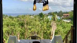 A dining area with a table and chairs, a hanging light fixture, and a view of a lush green landscape with trees and buildings in the distance. - Minutes from the beach, modern home with ocean views. Pet friendly!   (Rincón)