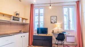Un espace de vie compact comprenant un coin cuisine, un téléviseur à écran plat, un bureau avec une chaise et une fenêtre avec des rideaux.