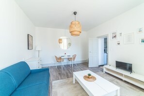 Un salon moderne avec un canapé bleu, une table basse blanche et un téléviseur posé sur un meuble blanc. On y trouve également un coin repas avec une table et des chaises, ainsi qu’un miroir accroché au mur.