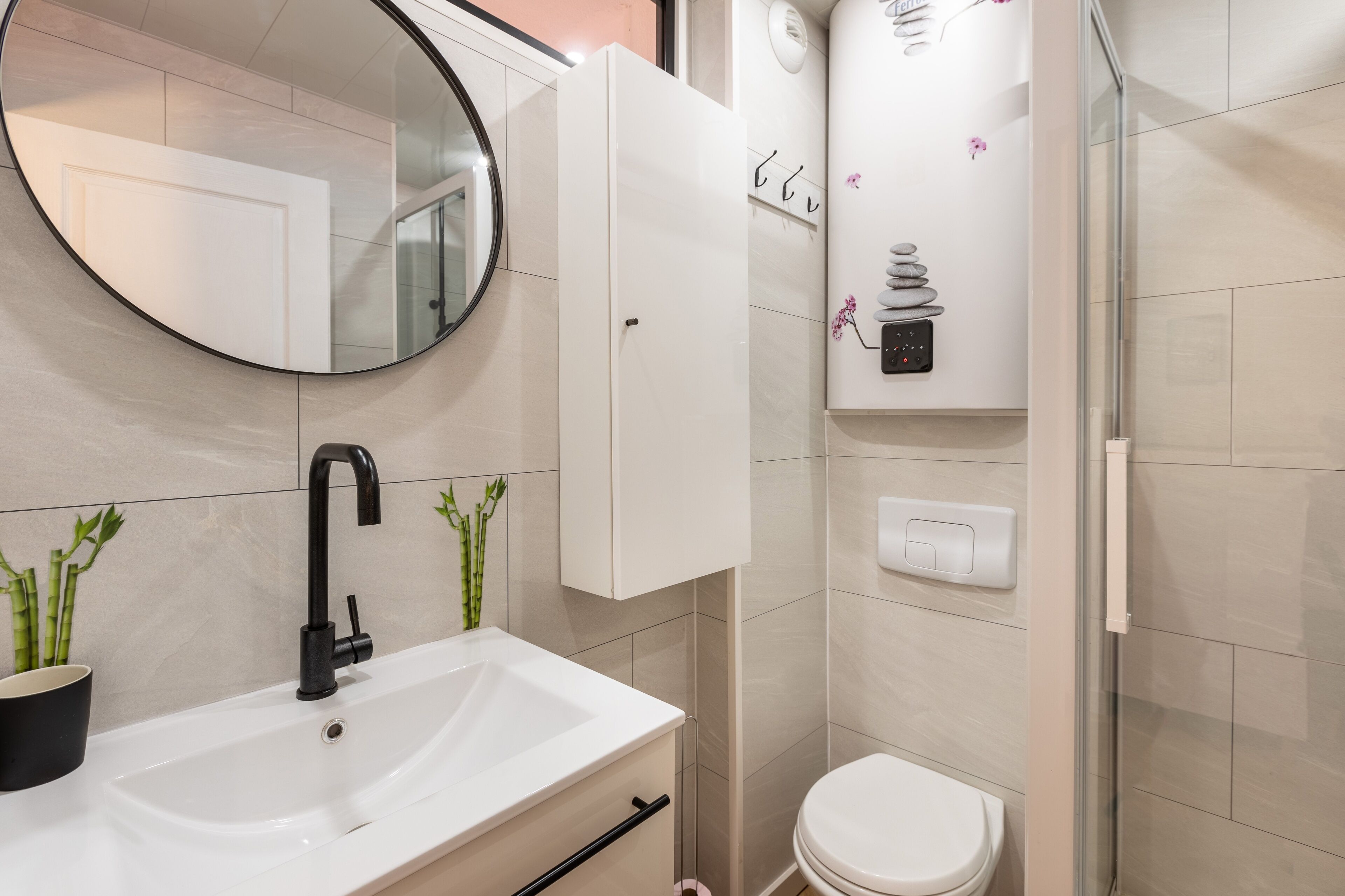 Un baño moderno con un lavamanos blanco, grifo negro y un espejo redondo. Hay un inodoro y una cabina de ducha con paredes de vidrio.