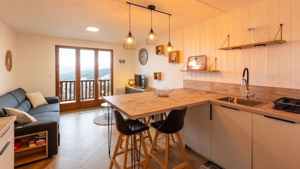 A modern kitchen with a wooden island, bar stools, pendant lights, and a large window overlooking a balcony.