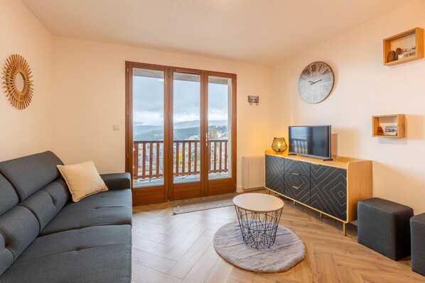 A modern living room with a grey sofa, a round coffee table, a TV on a wooden cabinet, and a balcony with a view.