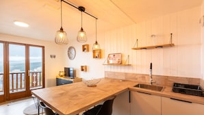 A modern kitchen with a wooden island, black chairs, pendant lights, and a view through large sliding doors.